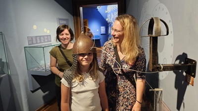 Zwei Frauen und ein Mädchen im Urgeschichte-Museum. Das Kind hat einen ungarischen Kriegshelm auf. Alle lachen