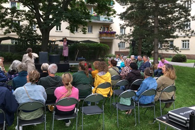 Publikum sitzt auf Klappsesseln im Hof eines Gemeindebaus in Wien und schaut sich das Utopia Theaterstück an.