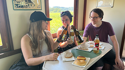 Zwei Frauen und ein Mann sitzen im Zug an einem Tisch und prosten sich mit Radler-Flaschen zu