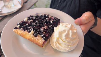 Mit einer Hand wird ein weißer Teller gehalten. Auf ihm befindet sich ein Stück Kuchen, ganz mit Heidelbeeren belegt. Daneben ein runder Gupf Schlagobers.