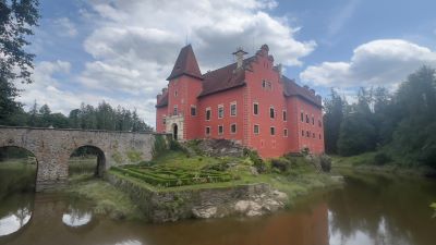 Blick über eine kleinen Teich auf einen im französischen Stil gestalteten kleinen Vorgarten mit einem zweigeschossigen Schloss mit roter Fassade. Über den Teich führt eine alte Brücke mit steinernen Gewölben zum Schlösschen. Der Teich ist von Nadelwäldern umgeben. Der Himmel blau mit ziehenden weißen Wolken.