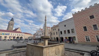 Ein Steinbrunnen mit quadratischem Becken inmitten eines mittelalterlichen Hauptplatzes. Man sieht sgrafittogeschmückte Bürgerhäuser und  den mit einem Spitzhelm gezierten Turm der Kostel Nanebevzetí Panny Marie, der Kirche der Himmelfahrt der Jungfrau Maria.