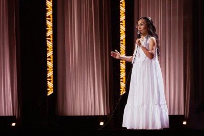 Zu sehen ist ein Moment aus der Show Single Lady von Ali Wong. Eine Nahaufnahme einer asiatischen Frau mit Brille auf der Bühne. Sie trägt ein weißes Kleid und hält ein Mikrofon in der Hand. Hinter ihr ist eine Art rosa Samthintergrund zu sehen.