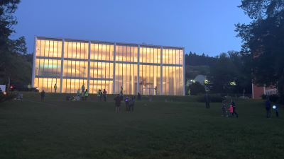 Das Bata Denkmal in Zlín: Ein vornehmlich aus Glas bestehendes Gebäude, streng in den Proportionen sechs zu drei errichtet. Von innen hell erleuchtet. Davor im Abendlicht einige Passanten, die um das Gebäude gehen beziehungsweise in der Wiese davor liegen.
