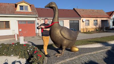 Ein Mann hält das rote Halsband einer mehr als mannshohen Metallstatue einer Gans, unter der ein Metallei liegt. Szene auf einem Dorfplatz mit einer Reihe ebenerdiger Häuser.