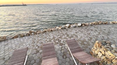 Drei Holzliegen stehen in der Abenddämmerung an einer steingepflasterten Uferpromenade mit Blick auf die Wasserfläches des Donau-Stausees Gabčíkovo–Nagymaros.