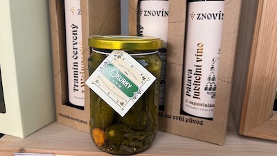 Ein Gurkenglas, mit der Aufschrift Okurky, tschechisch Gurken. Dahinter in Kartonboxen verpackte Weinflaschen mit weißem Etikett.