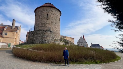 Eine romanische runde Rotunde, etwas erhöht auf einer begrünten Anhöhe. Daneben in zweistöckiges Gebäude mit hohem Rauchfang. Dahinter eine Kirche und Kapelle. Davor ein stehender Mann.
